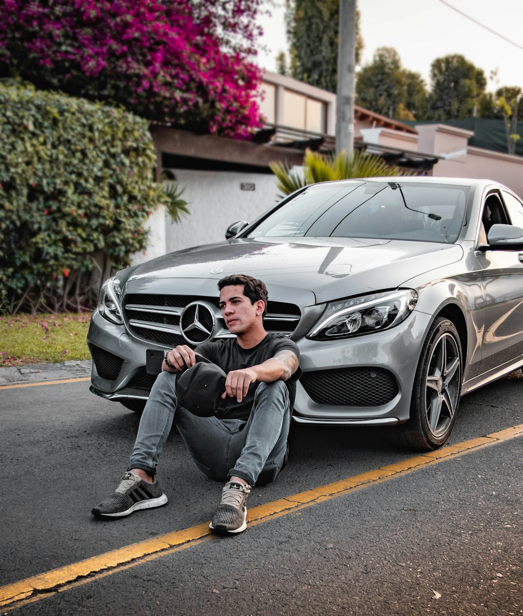 unknown celebrity sitting siting beside silver Mercedes-Benz car outdoors thinking about auto insurance questions